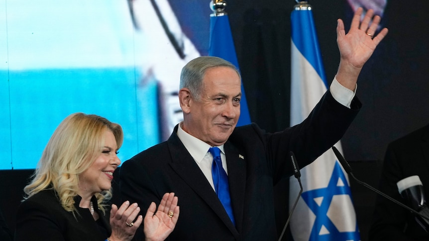 A suited man waves from a stage while standing next to a woman and Israeli flags.