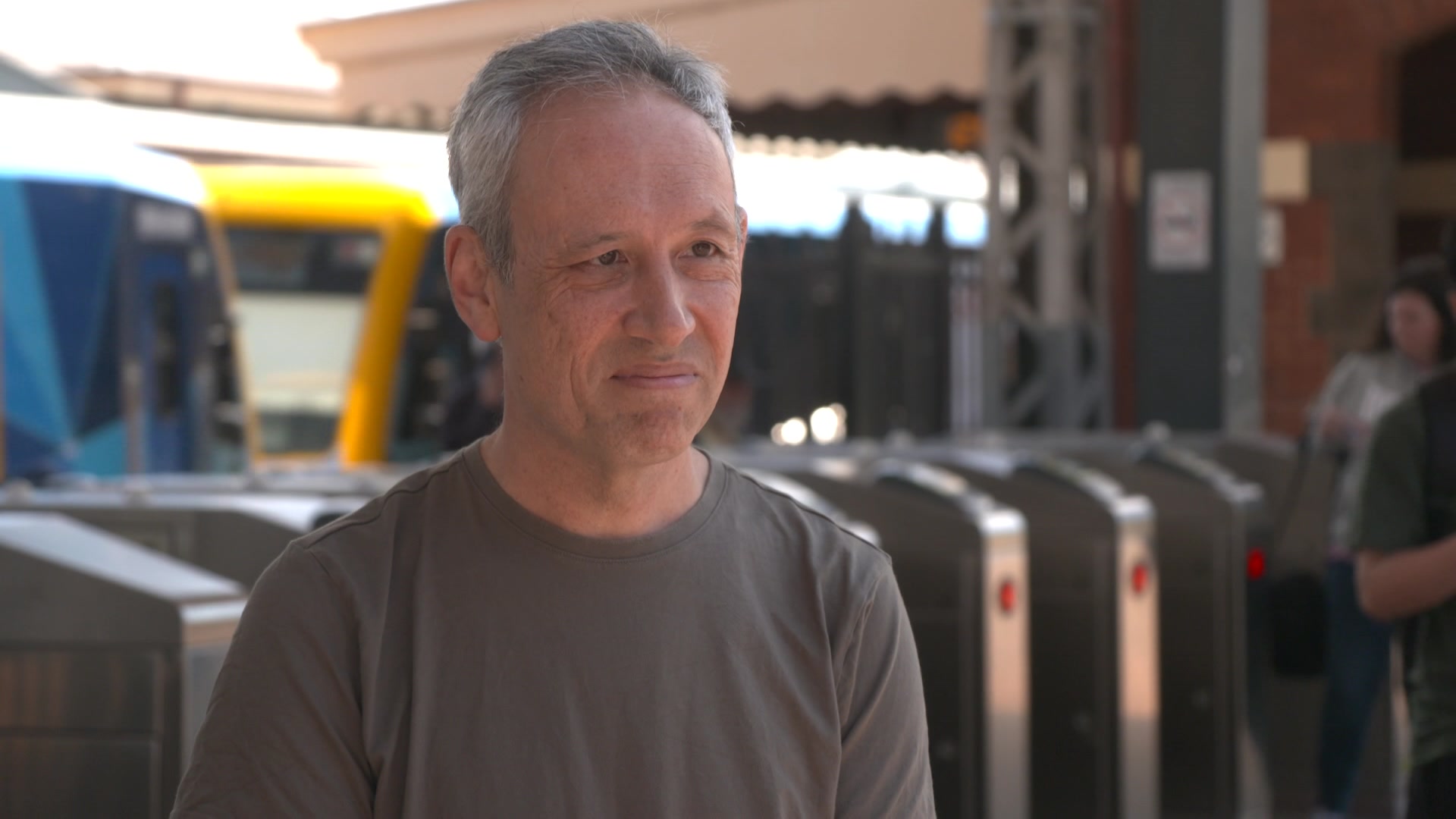 Daniel Bowen wears a grey t-shirt and stands in front of entry turnstiles to a railway station with a train in the background.