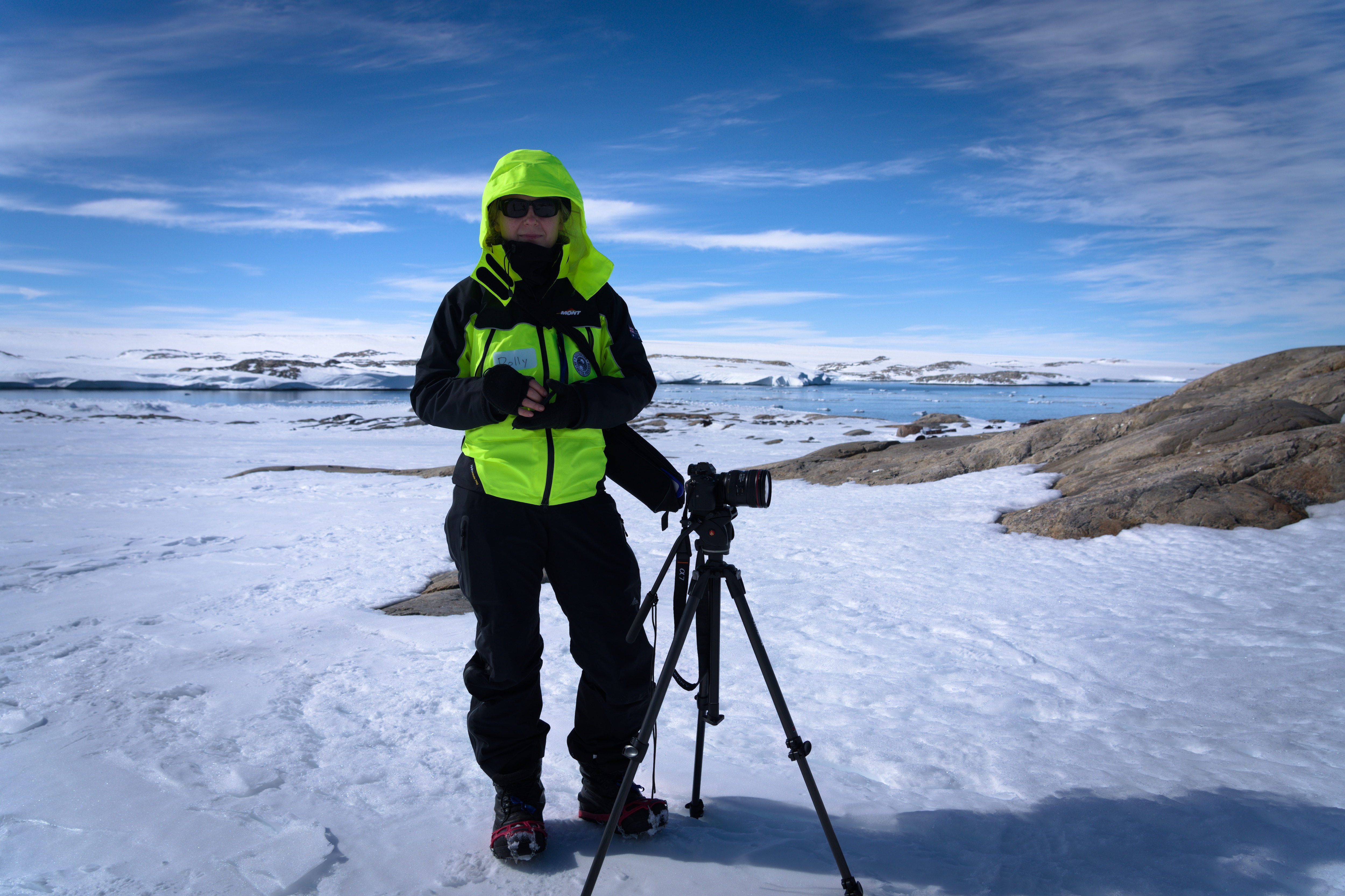 Polly Stanton usa equipamento de neve preto e amarelo fluorescente e fica ao lado de uma câmera em um tripé no gelo da Antártica.