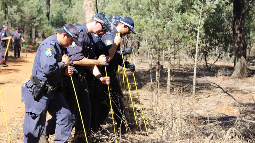 Uniformed police officers stand in a line probing the ground with a yellow stick with every step they take