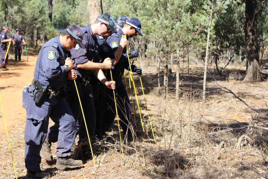 Uniformed police officers stand in a line probing the ground with a yellow stick with every step they take