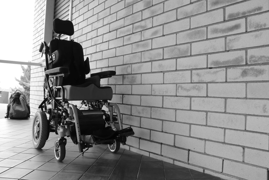 A black wheelchair is seen next to a brick wall. There is a window behind it, with a backpack leaning on it.