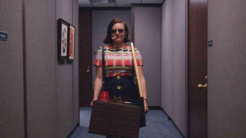 Mad Men's Peggy Olson, played by Elisabeth Moss, walks down a hallway carrying a box while smoking a cigarette.