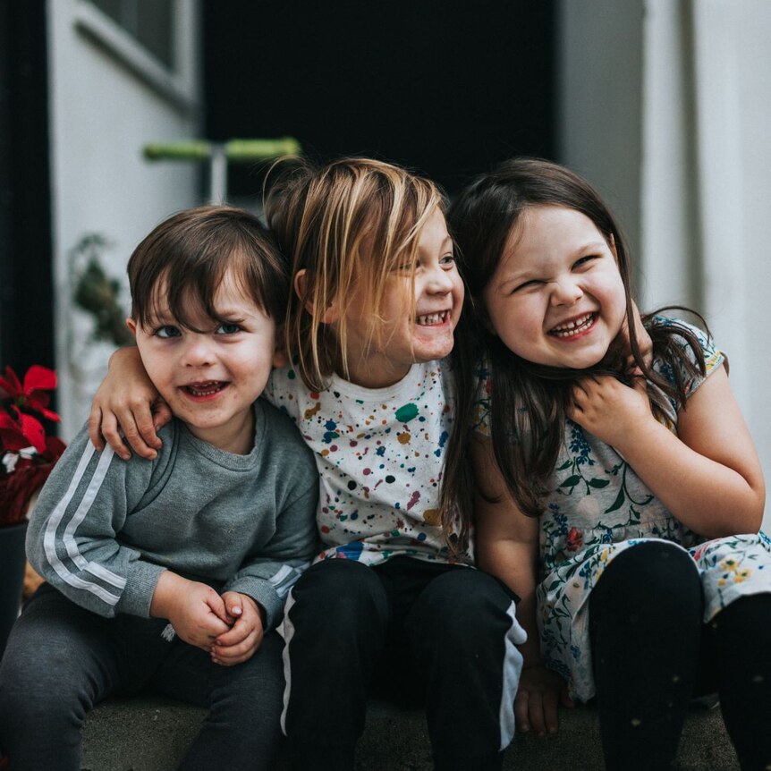 three small children sitting on a step with their arms around each other smiling and looking at the camera