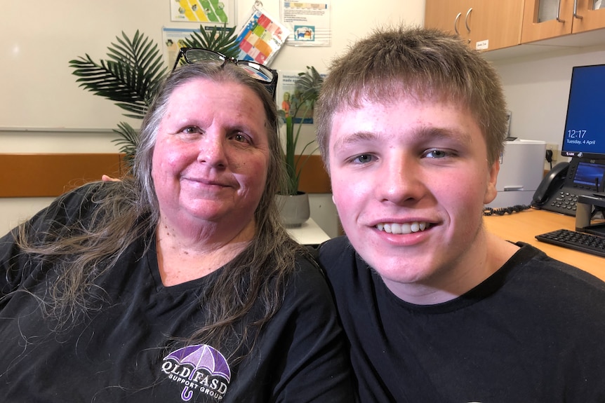 A mother and son sitting in an office. A computer, small palm tree and some notes pinned to the wall are in the background. 