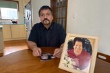 Spiro Vasilakis sitting at a wooden table next to a smiling photo of his late mother.
