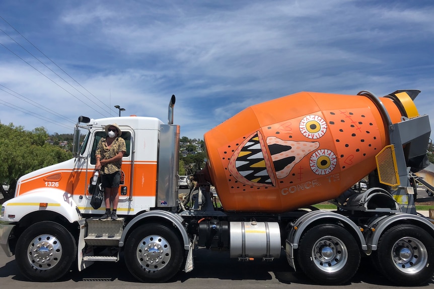 A man standing on an orange cement truck with a scary face drawn on its side