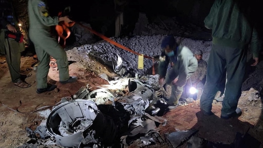 People in military garb stand around a hole in the ground littered with parts of metal and lit by portable lights