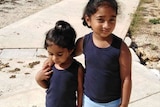 Kopika and Tharnicaa in blue navy tanks, light blue leggings and black crocs standing on a path, building and fences behind.