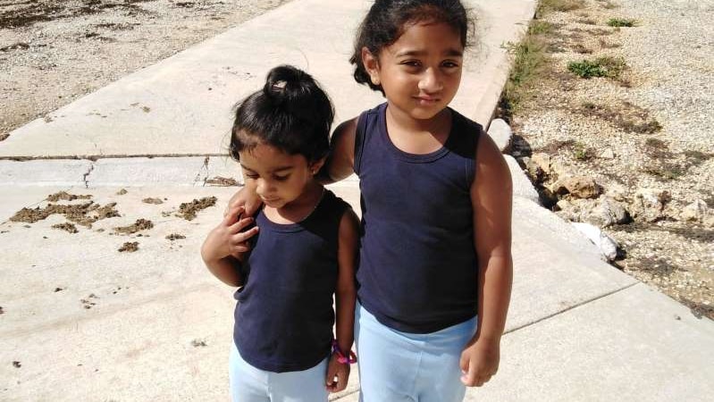 Kopika and Tharnicaa in blue navy tanks, light blue leggings and black crocs standing on a path, building and fences behind.