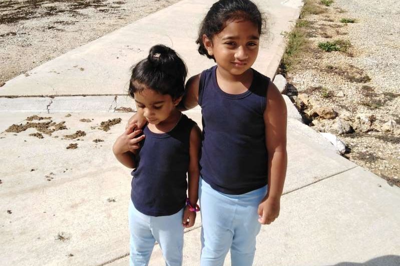 Kopika and Tharnicaa in blue navy tanks, light blue leggings and black crocs standing on a path, building and fences behind.