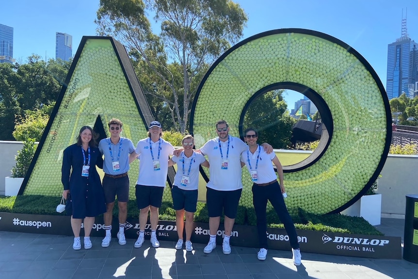 Six people stand arm in arm, smiling at the camera, in front of large "AO" initials symbolising the Australian Open tennis.