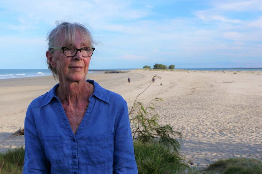 A white woman in her 60s, wearing a blue linen shirt, sitting in front of a sandy stretch with an off-leash dog behind her