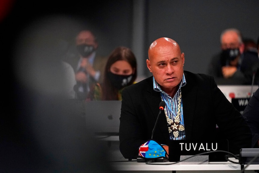 A man sits behind a Tuvalu name plate at a desk at COP-26, with a pensive expression.