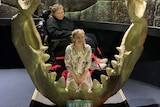 A young girl sits in the centre of the jaws of a large shark with her sister in a wheelchair behind her
