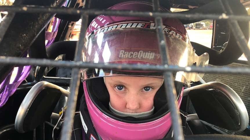 A young girl in a purple helmet peers through the protective bars of a speedway vehicle.