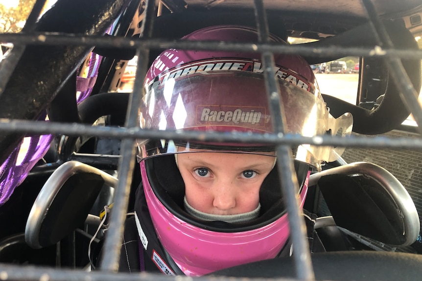 A young girl in a purple helmet peers through the protective bars of a speedway vehicle.