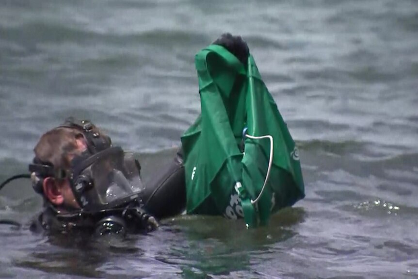 police diver hands a green bag full of cables from the lake surface to a waiting officer