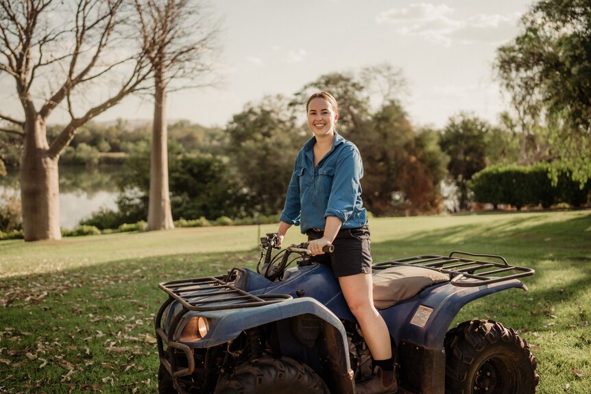 A woman wearing a blue shirt and shorts stands up on a quad bike 
