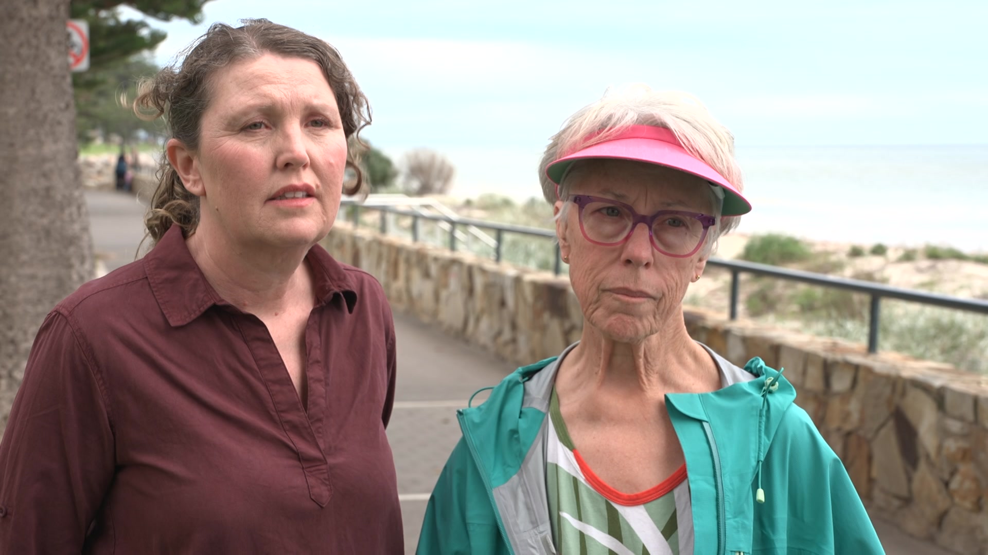 Two women look upset at the camera while standing on a footpath next to sand dunes and a beach