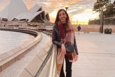 A woman stands before the Sydney Opera House and a sunset smiling