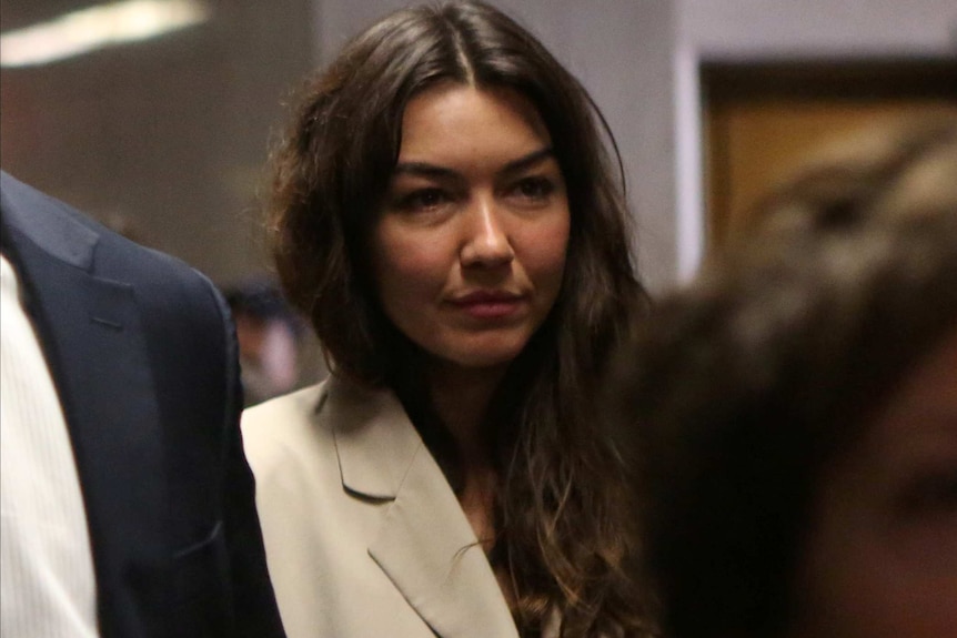 A woman with long hair, wearing  a cream jacket is flanked by two men (obscured) in a hallway.