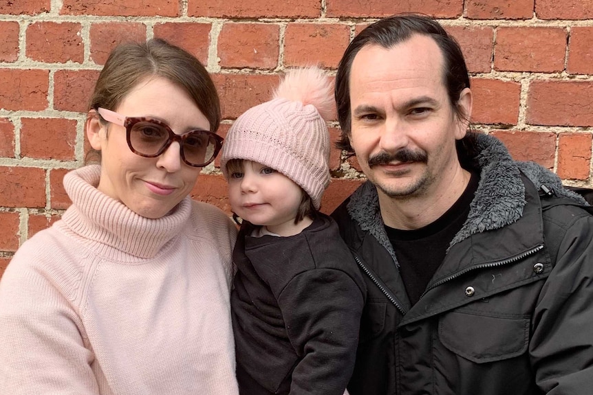 Shannon, Isla and Chris Pierucci stand together in front a red brick wall. They wear winter attire.