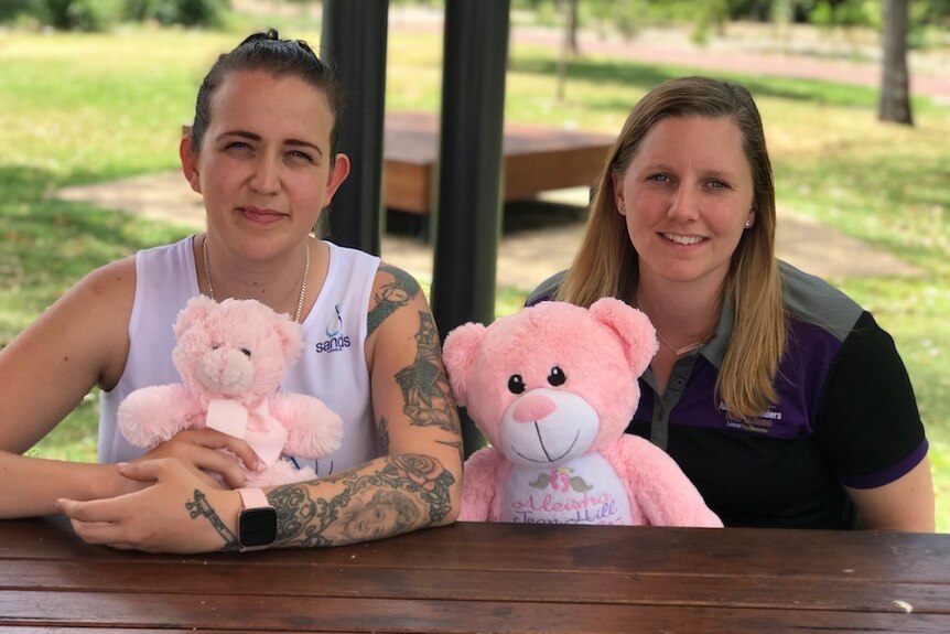 Two women, both holding pink teddy bears, sit at a picnic bench and look into the camera