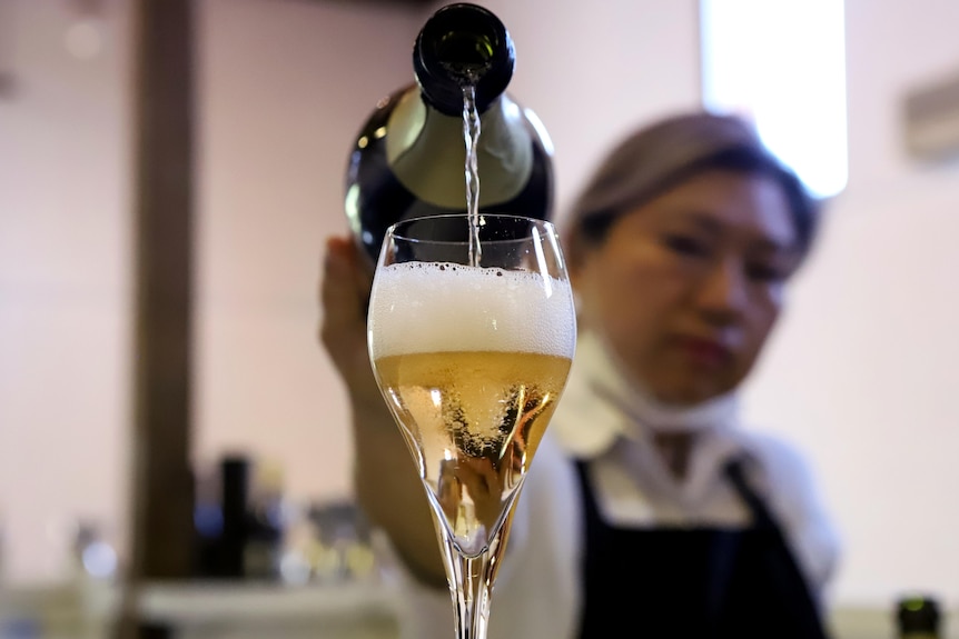 Woman wearing white shirt and black apron pours sparkling wine into a champagne flute