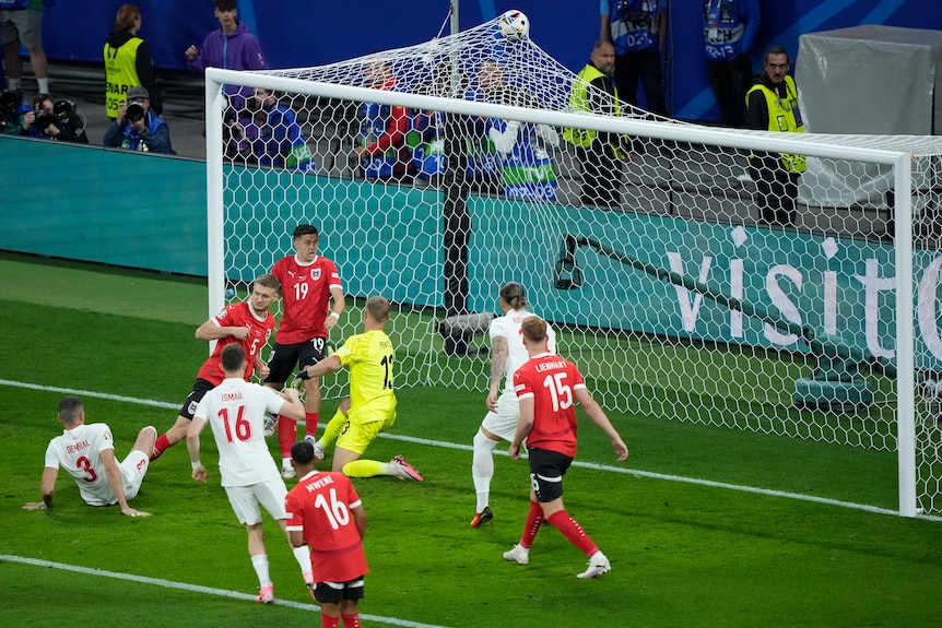 A group of Türkiye and Austria footballers stand around, as the ball hits the top of the net - the goalscorer sits on the turf.