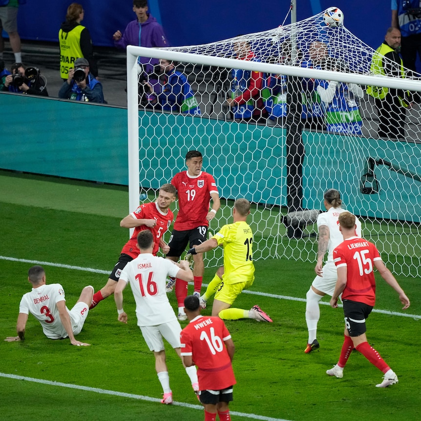 A group of Türkiye and Austria footballers stand around, as the ball hits the top of the net - the goalscorer sits on the turf.