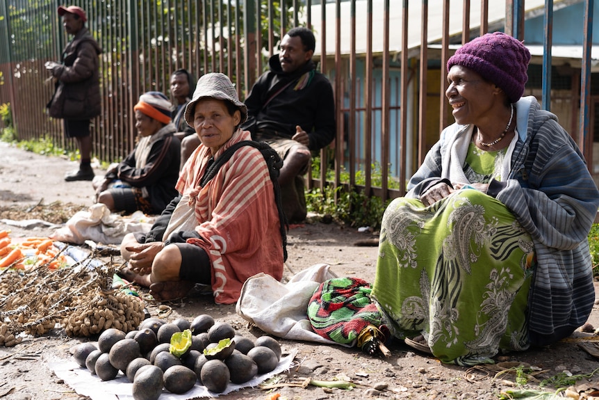 Three older women sitting on a dirt road selling produce. 