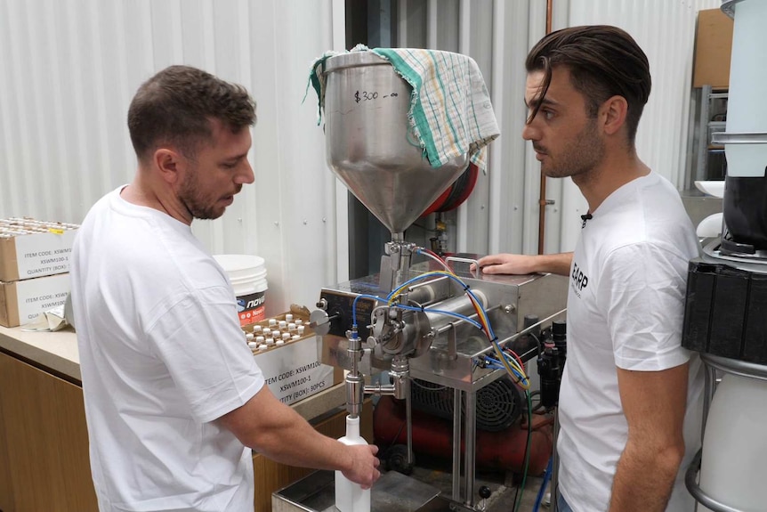 Two young men, wearing white t-shirts and sporting rather trendy haircuts, operate a still.