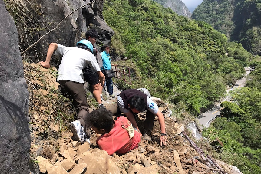 A tourist wearing a red shirt is pulled from rubble by rescue workers wearing helmets on the side of a mountain.