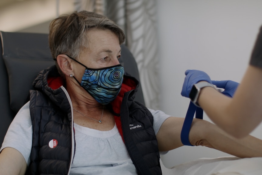 A woman sits on a hospital chair in a facemask, awaiting a blood test.