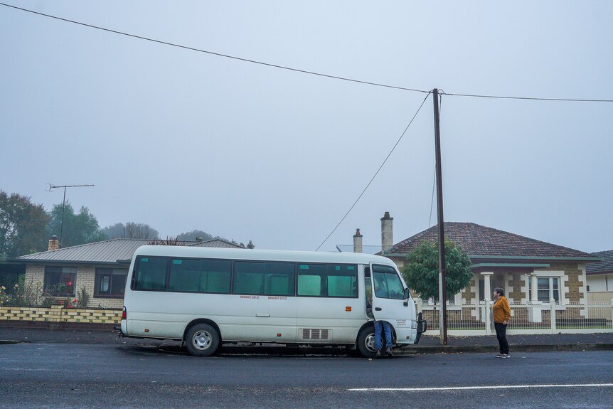 A man and woman stand next to a white mini bus in a suburban street on a foggy morning.