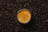 A cup of espresso sits on a surface, surrounded by dark brown coffee beans.