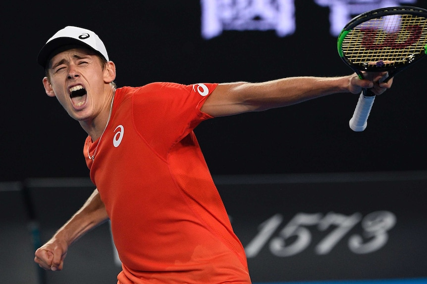 Alex de Minaur, in a red shirt, pumps his fist and screams in delight after winning a tennis match.