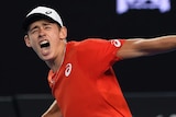 Alex de Minaur, in a red shirt, pumps his fist and screams in delight after winning a tennis match.