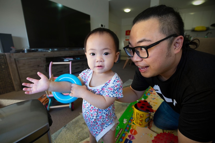 A young baby girl smiles at the camera while her dad supports her back and smiles.