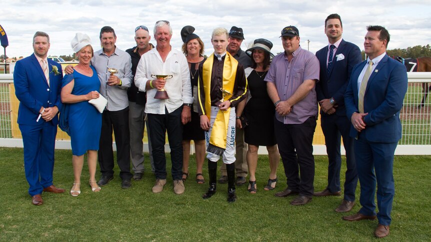 A dozen people stand in front of a horse racing course with two trophies.