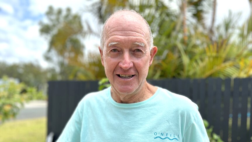A man with white hair and a light blue t-shirt smiles at the camera on a sunny day.