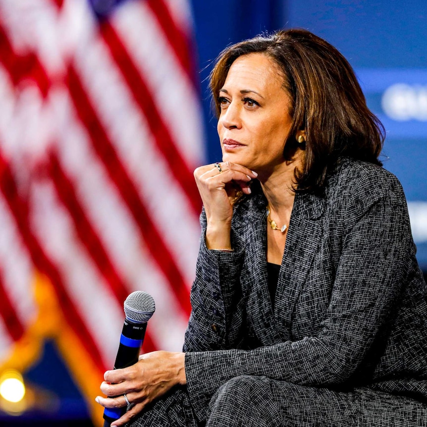 Kamala Harris in front of a row of US flags resting her head on her chin