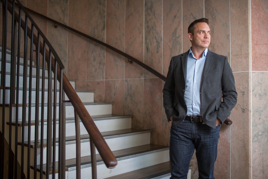 A man stands with his hands in his pockets, at the bottom of a curved staircase.