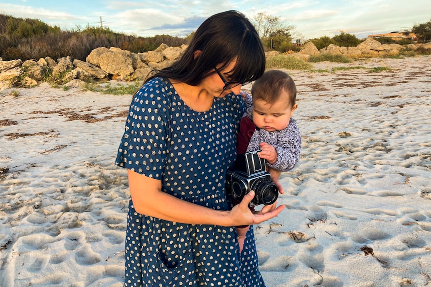 A woman with dark hair holds a dark-haired baby on the beach, as they both look at a camera in the woman's hand