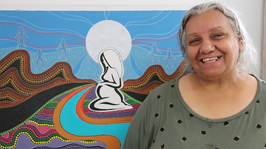 Woman smiling on right in front of indigenous dot painting of swirls and pregnant woman