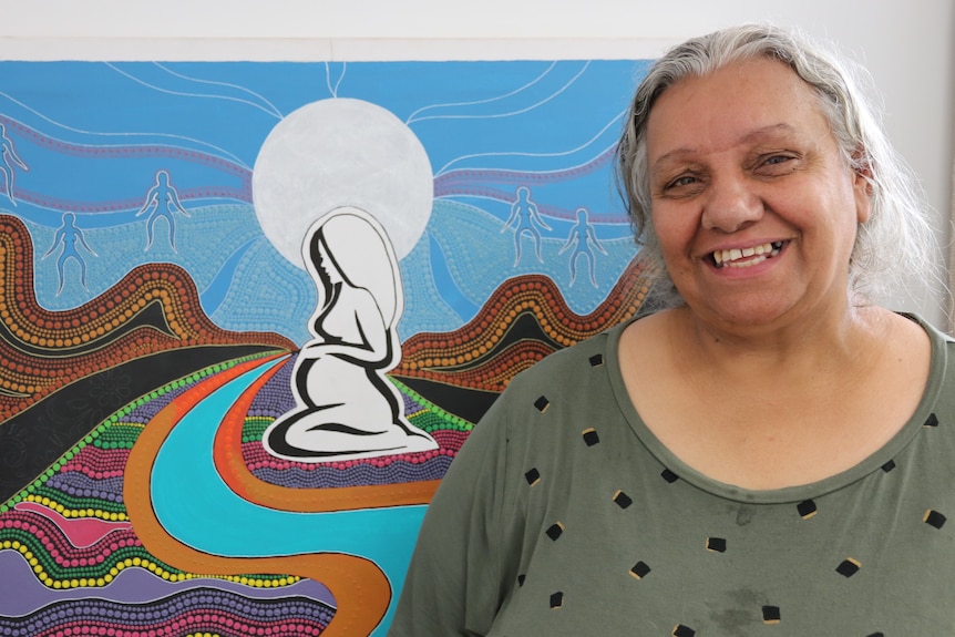 Woman smiling on right in front of indigenous dot painting of swirls and pregnant woman