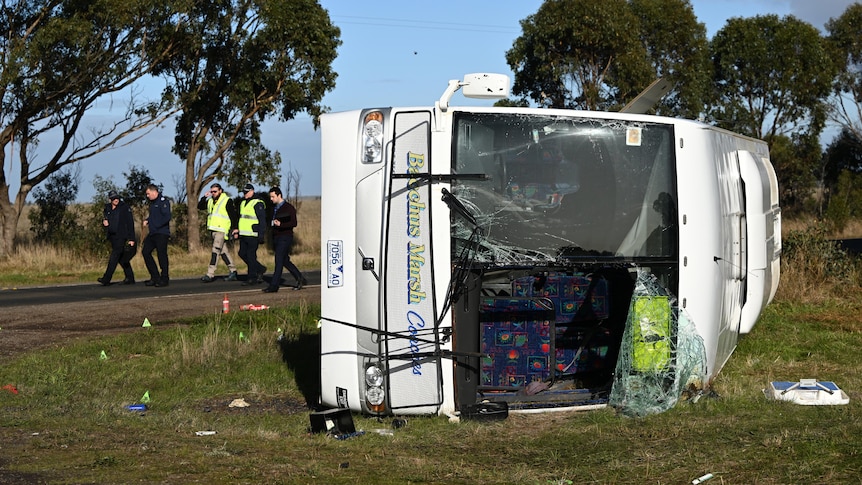 A school bus rolled onto its side with the impact of a crash visible, with emergency services walking behind.