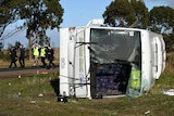 A school bus rolled onto its side with the impact of a crash visible, with emergency services walking behind.