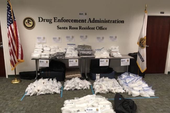 Piles of drugs packaged in individual plastic bags sit on the floor and tables in a drug enforcement agency office.